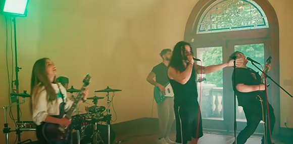 A musical band performs indoors with a guitarist, drummer, bassist, singer, and another vocalist.