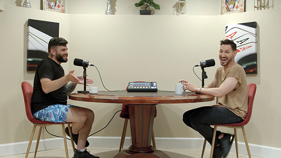 Two men sit across from each other at a round wooden table with microphones and a mixer between them.