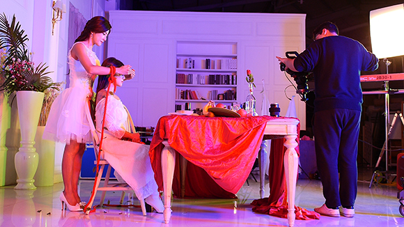A photographer films a model seated at a table draped in red cloth during a studio photoshoot.