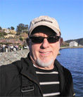A man wearing a tan cap and sunglasses smiles in front of a blue ocean and coastal hills.