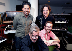 Four men smiling in a music studio filled with various electronic keyboards and audio equipment.