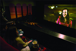 Two people sit in a dark movie theater watching a woman smiling on a large screen.