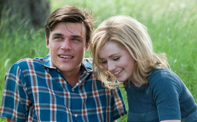 A man in a blue plaid shirt and a blonde woman in a grey shirt smile while sitting in a grassy field.