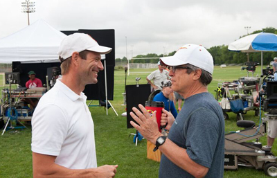 A man in a white polo and cap talks to a man in a blue shirt and white cap on a grassy film set.