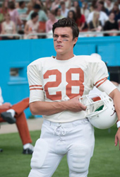 A man in a white and orange number 28 football jersey holds a white helmet on a green field.