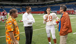A football player in white and orange uniform talks with three men on a green indoor stadium field.