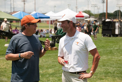 Two men in casual attire stand on a grass field talking near event tents.