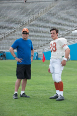 A man in a blue shirt and shorts stands next to a football player in a white jersey on a grass field.
