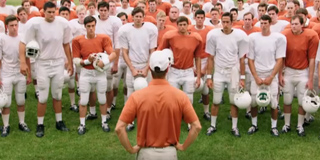 A football team in white and orange uniforms stands on a field facing a coach in an orange shirt.