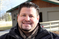 A smiling man with dark hair and a goatee wearing a black jacket and a patterned scarf.