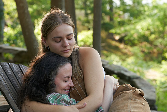 A young woman with blonde hair hugs an older woman with dark hair in a wooded area.