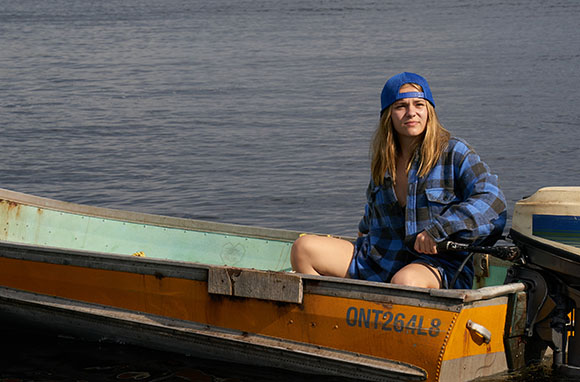 A woman wearing a blue cap and plaid shirt sits in a yellow and green motorboat on the water.