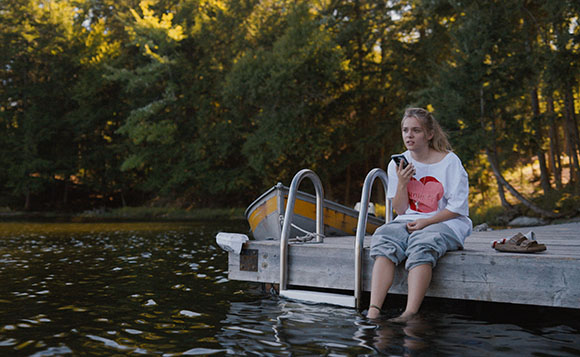 A young woman in a white t-shirt sits on a wooden dock with her feet in the water, holding a phone.