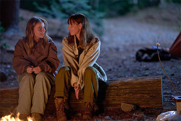 Two women sit together on a large log near a campfire in a wooded area at night.
