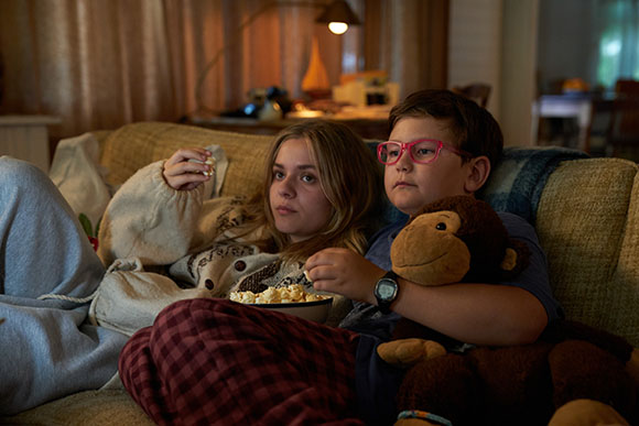 Two children sit on a couch, one holding a bowl of popcorn and the other holding a brown stuffed monkey.
