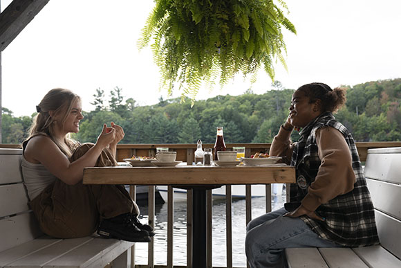 Two people sit at a wooden table on a deck overlooking a lake and forested hills.