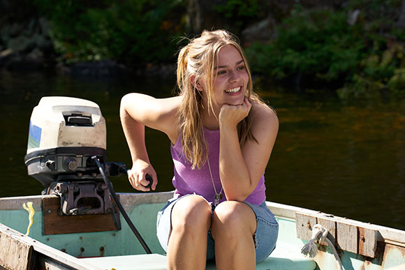 A smiling young woman with blonde hair sits on the edge of a small boat on a lake.