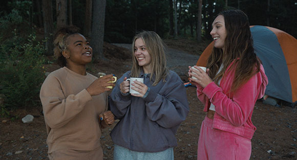 Three young women stand together outdoors at dusk, smiling and holding mugs near a tent.