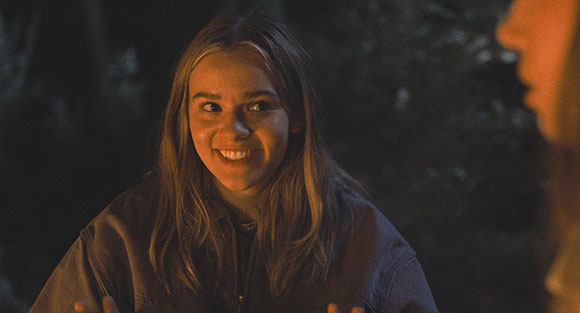 A smiling young woman with blonde hair looks toward a person in a dark, outdoor setting.