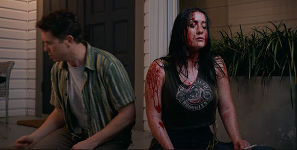 A man in a striped shirt sits next to a woman covered in blood outside a house at night.