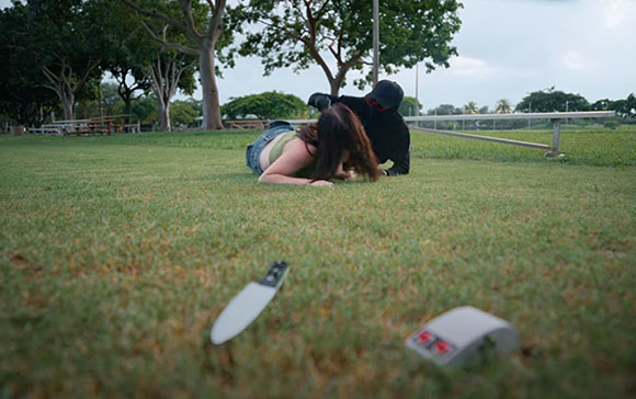 A person in black leans over a person lying face down on a grassy field near a knife on the ground.