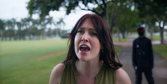 A woman with reddish-brown hair speaks toward the camera in a park with a blurred figure behind her.