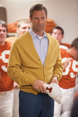 A man in a yellow cardigan and blue shirt stands before football players in orange jerseys.