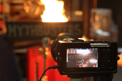 A video camera on a tripod records a bright fire and a sign that reads MYTHBUSTERS in a dark setting.