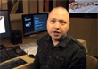 A man in a dark shirt sits in front of computer monitors in a studio setting.