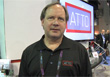 A man in a black polo shirt stands in front of a large blue and red sign that reads ATTO.