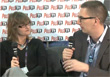 A woman and a man sit facing each other holding microphones against a branded backdrop.