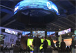 A large, glowing blue circular screen suspended above a dark, busy indoor hall with people below.