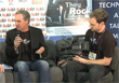 A man in a black blazer speaks into a microphone while another man sits beside him holding a camera.