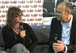 A woman in a black shirt and a man in a suit sit on a couch holding microphones during an interview.