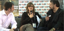 A woman in glasses speaks into a microphone while seated between two men during an interview.