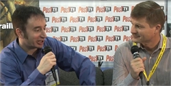 Two men sitting side by side, holding microphones and looking at each other against a branded backdrop.