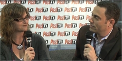 A woman with glasses and a man in a blazer hold microphones while looking at each other against a Post TV logo wall.