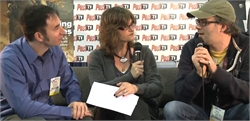 A man in a blue shirt, a woman with glasses holding a microphone, and a man in a cap talking.