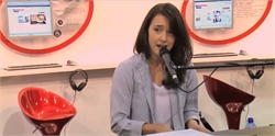A woman with dark hair wearing a light blue shirt speaks into a microphone in a studio.
