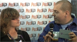 A man in a blue shirt holds a microphone and a small device while speaking to a woman with glasses.