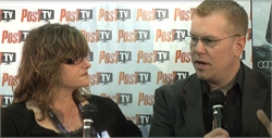 A woman with wavy hair and glasses looks at a man in a suit speaking into a microphone.