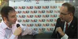 Two men sit facing each other, holding microphones against a backdrop of logos.