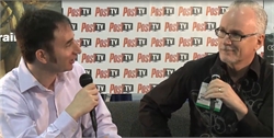 A man in a white shirt holds a microphone while interviewing a man in a black patterned shirt.