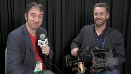 Two men sit before a black curtain; one holds a microphone and the other holds a large professional camera.
