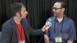 A man in a dark blazer holds a microphone toward a man in a blue shirt during an interview.
