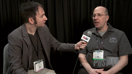 A man in a grey blazer holds a microphone toward a man in a grey polo shirt during an interview.