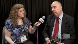 A woman with curly hair holds a microphone toward a man in a suit and red tie.
