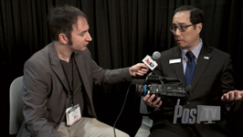 A man in a grey blazer holds a microphone toward a man in a suit during an interview.
