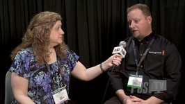 A woman holds a microphone toward a man during an interview against a dark curtain background.
