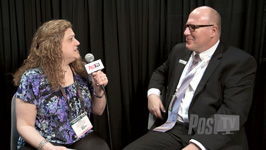 A woman holding a microphone interviews a man in a suit against a black curtain background.